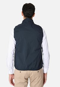Gilet senza maniche, leggero e navy, con colletto alto; texture liscia e orlo elastico; indossato sopra una camicia a maniche lunghe bianca.