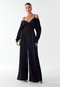 Moteris vilki juodą šoninį šuoliuką su plačiomis kojomis, laiko mažą metalinį delninuką ir dėvi auksinius auskarus bei šlepetes.