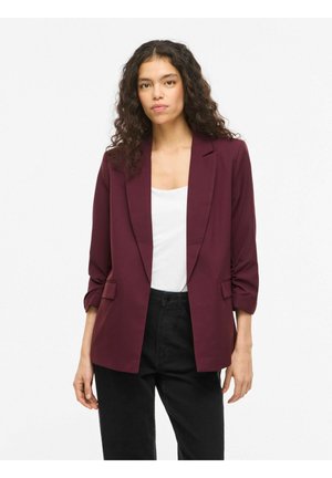 Kvinde med krøllet hår iført en burgunder blazer, hvid top og sorte bukser, stående mod en ensartet hvid baggrund.