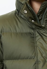 Olijfgroene pufferjas met een hoge kraag, drukknoopsluiting en een getextureerde ritssluiting. Heeft een doorgestikt patroon over het materiaal.