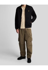 Svart zip-up jacka med ståkrage och gult märke, i kombination med beige polo och olivgröna cargobyxor. Texturerade svarta skor.