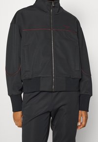Giacca nera con zip e alto collo, cuciture rosse a contrasto e tasche laterali. Realizzata in un tessuto liscio; presenta orlo e polsini a costine.