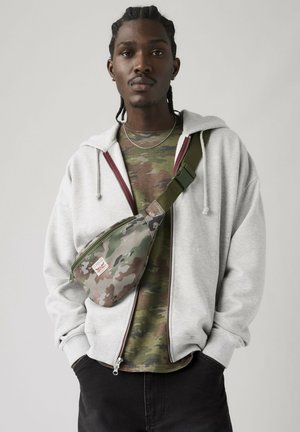 Young man with braided hair wears gray zip-up hoodie, camouflage shirt, crossbody camo bag, and dark pants, standing against plain background.