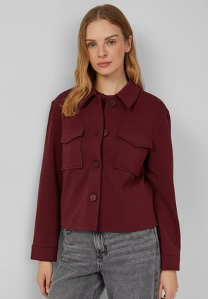 Veste boutonnée courte bordeaux en matériau doux, dotée de deux poches poitrine, d'un col pointu et de manches longues. Détail de tissu texturé.