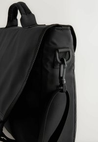 Fekete vászon üzenettáska vállpánttal, amely műanyag kapoccsal van rögzítve és párnázott fogantyúval, egyszerű háttér előtt.