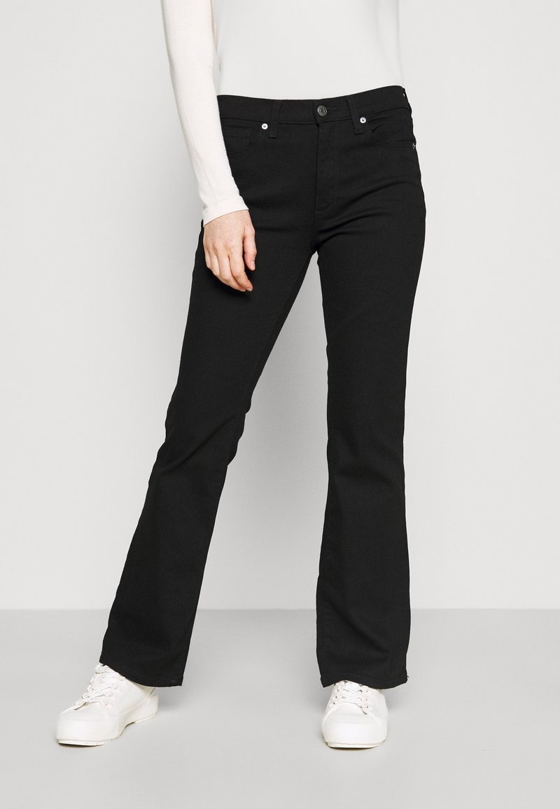 Jean noir évasé en denim, avec une fermeture à bouton et des passants de ceinture, associé à des baskets blanches sur un fond neutre.