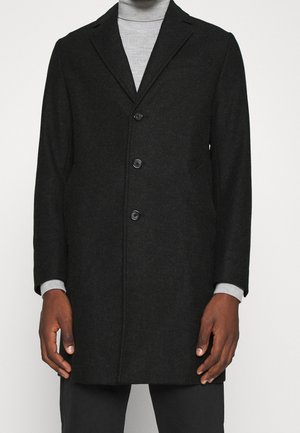 Un homme porte un manteau en laine noir boutonné jusqu’aux genoux, par-dessus un col roulé gris clair et un pantalon noir, debout devant un fond uni.