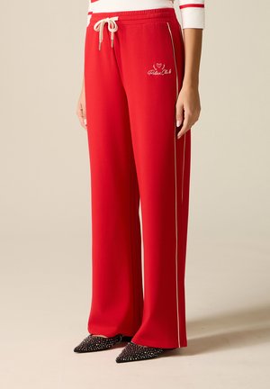 Pantaloni rossi a gamba larga con piping bianco laterale e scritta "Relax Club", indossati con scarpe nere con borchie e punta a punta.