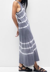 Šedé tie-dye maxi šaty bez rukávů s bílými kruhovými vzory, vlající materiál a uvolněná silueta doplněné o splétané černé sandály.