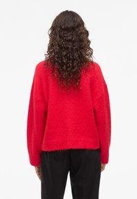 Pull en maille rouge avec une texture douce, coupe ample et manches larges. Présente une longueur courte et un ourlet côtelé. Sans motifs ni éléments en métal visibles.