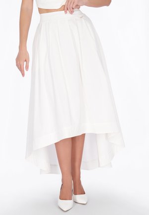 Femme portant une jupe midi blanche à ourlet asymétrique et des talons blancs à bout pointu, debout devant un fond blanc.