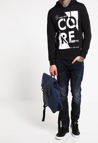 Sudadera negra con estampado gráfico blanco, jeans azules, zapatillas negras y una bolsa azul oscura con tapa y correa para el hombro. Atuendo casual.