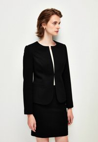Černý strukturovaný blazer s kulatým výstřihem, vybavený skrytým předním zapínáním a dlouhými rukávy, v kombinaci s odpovídající černou sukní.