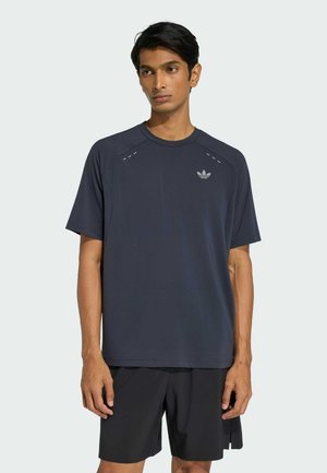 Homme portant un t-shirt Adidas bleu marine foncé à manches courtes et un short noir, debout avec les bras détendus contre un fond gris clair.