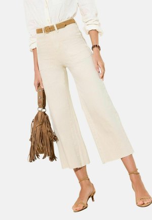 Person iført cremefarvede vidde bukser med høj talje, hvid langærmet skjorte, tan bælte, tan ankelremme hæle og holder en tan frynset taske.