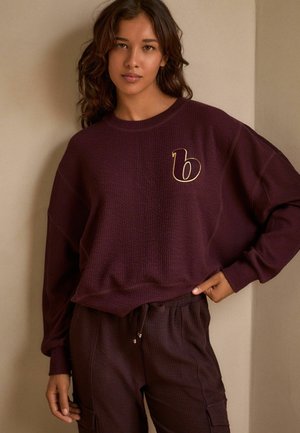 Felpa bordeaux con texture, dotata di scollatura tonda e accentuata da una lettera "b" dorata sul petto, abbinata a joggers con coulisse dello stesso colore.