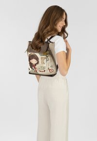 Beiger Rucksack aus Stoff mit einer verspielten Charakterillustration, dunkelbraunen Akzenten, Reißverschluss und verstellbaren Schulterriemen.