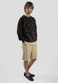 Pull marron avec motif ondulé noir, short clair et mocassins noirs. Coupe décontractée, tissu texturé et logo visible sur le pull.