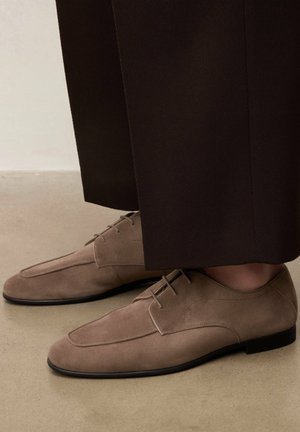 Chaussures à lacets en daim marron portées avec un pantalon marron foncé, posées sur un sol beige.