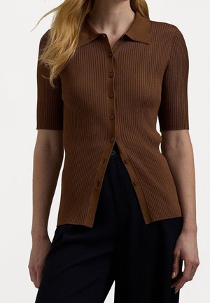 Mujer con un top marrón ajustado de punto acanalado, de manga corta y con botones, y pantalones negros sueltos, de pie con los brazos relajados a los lados.