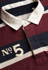Polo rayé bordeaux, marine et blanc avec un col blanc. Présente un "No. 5" brodé et une fermeture à boutons sur la patte de boutonnage. Texture en coton doux.