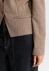 Brown and cream checked blazer with a structured fit, single pocket, and button closure, paired with dark denim jeans.