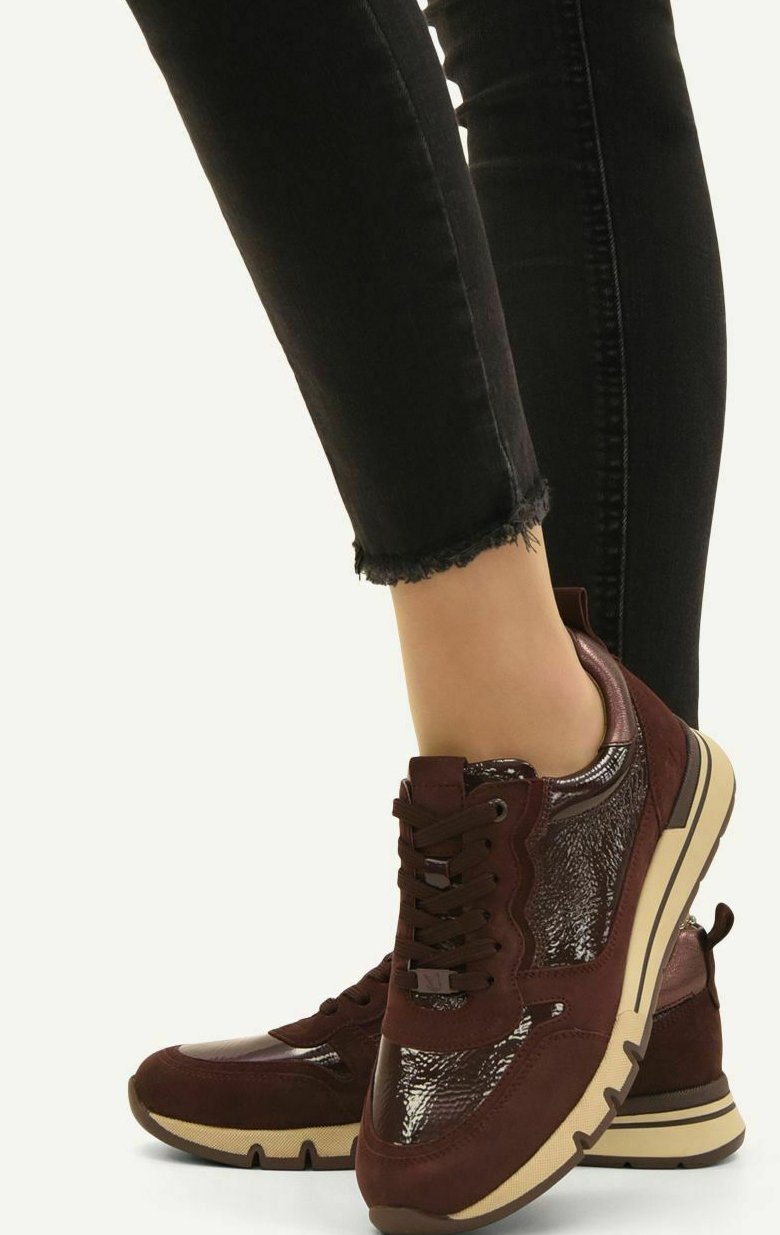 Pieds portant des baskets rouge foncé avec des semelles beige et des jeans skinny noirs aux ourlets effilochés, posés sur un fond uni.