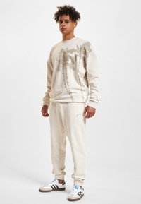 Béžová mikina s grafikou palmového stromu, doplněná o odpovídající tepláky s logem. Bílé sneakers s černými pruhy dotvářejí celkový vzhled.