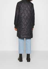 Cappotto lungo nero imbottito con colletto alto, caratterizzato da un motivo a diamante, maniche lunghe e polsini a coste, abbinato a jeans azzurri chiari e stivali neri.