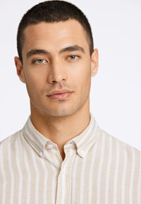 Camicia button-up beige con righe verticali bianche. Presenta un colletto e bottoni, realizzata in un tessuto leggero con una consistenza morbida.