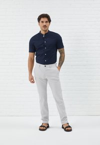 Chemise à manches courtes marine avec boutons, pantalon gris ajusté et sandales noires. L'arrière-plan présente un mur en briques blanches.