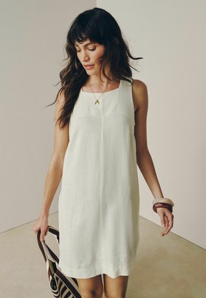 Hosszú, sötét hajú nő ujjatlan fehér ruhában és karkötőkben, csíkos táskát tartva, lefele néz egy egyszerű háttér előtt.