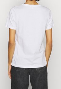 Bílý bavlněný tričko s krátkými rukávy a kulatým výstřihem, zobrazené zezadu. Nošeno s tmavými džínami, minimalistický design.