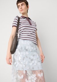 Polo shirt rayé en marron foncé et blanc ; associé à une jupe sheer bleu clair ornée de sequins floraux ; portant un sac à épaule marron.