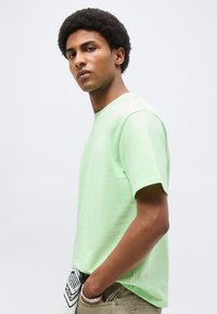 Νέος άνδρας με σγουρά μαλλιά, φορώντας ανοιχτό πράσινο μπλουζάκι και μπεζ παντελόνια, στέκεται με το ένα χέρι στην τσέπη μπροστά σε μονόχρωμο φόντο.