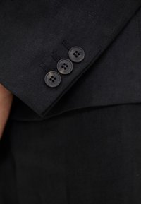Gros plan d'une manche de costume noire montrant trois boutons noirs alignés près du poignet sur un tissu texturé.