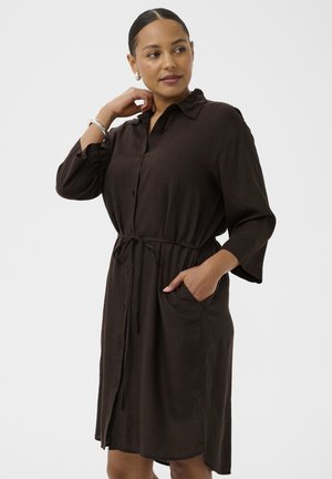 Kobieta w ciemnobrązowej sukience zapinanej na guziki z rękawami do łokcia, z paskiem w talii i jedną ręką w kieszeni, patrząca w bok, z włosami zaczesanymi do tyłu.