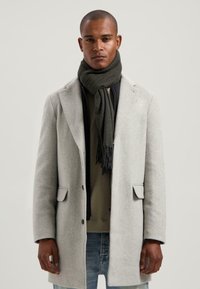 Grijze wollen jas met een getailleerd ontwerp, voorzien van twee vo pockets en een zwarte sjaal om de hals gewikkeld. Spijkerbroek maakt de outfit compleet.