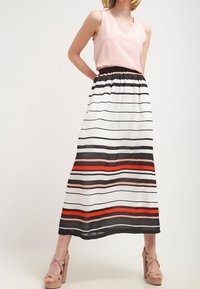 Jupe longue rayée en blanc, noir et rouge, avec une ceinture noire ; associée à un haut rose sans manches et des sandales compensées beiges.