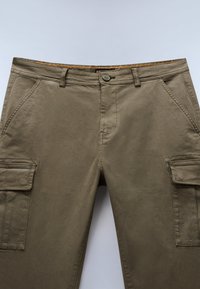 Cargo bukser i khaki grøn, lavet af blød bomuld. Har flere lommer, en frontlukning med knap og en dekorativ orange søm på taljebåndet.