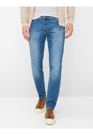 Man wearing light blue slim-fit jeans, beige zip-up jacket, white shirt, and brown lace-up sneakers, standing against white background.