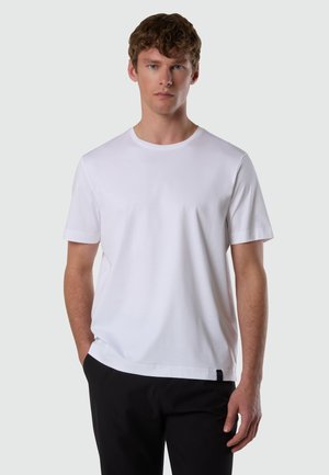 Giovane uomo dalla pelle chiara e capelli castani ricci, indossa una maglietta bianca a maniche corte semplice e pantaloni neri, in piedi con le mani rilassate.