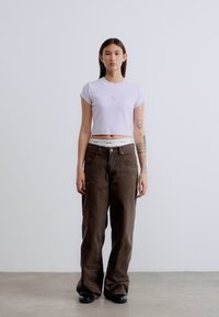 Ανοιχτό μωβ κοντομάνικο μπλουζάκι με μικρό λογότυπο, σε συνδυασμό με φαρδές καφέ παντελόνια και μαύρα παπούτσια. Απλός σχεδιασμός και ουδέτερα χρώματα.