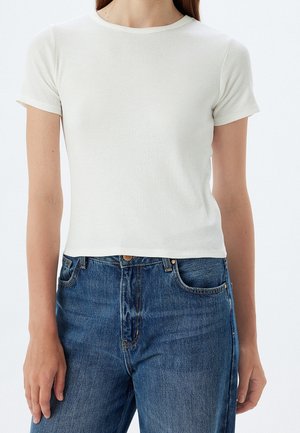 Kobieta ubrana w dopasowany prążkowany biały T-shirt z krótkim rękawem, połączony z niebieskimi wysokimi jeansami, stojąca na jednolitym tle.
