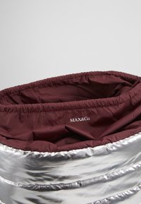 Srebrna pufasta torba z marszczonym bordowym wnętrzem. Posiada logo "MAX&Co." nadrukowane na krawędzi. Odbijająca, gładka faktura powierzchni.