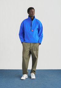 Μπλε μπουφάν με φερμουάρ μέχρι τη μέση, με ελαστικές μανσέτες και μπροστινή τσέπη, συνδυασμένο με πράσινο παντελόνι φαρδιάς γραμμής και λευκά αθλητικά παπούτσια με πολύχρωμες λεπτομέρειες.