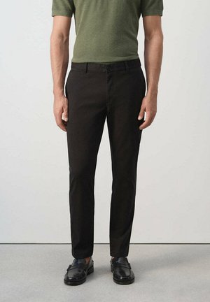 Hombre que lleva pantalones negros ajustados, zapatos de vestir negros y una camisa de manga corta verde oliva, de pie frente a un fondo liso.