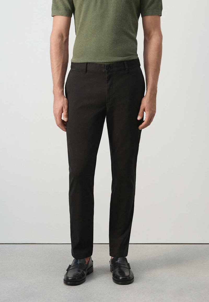Homme portant un pantalon noir slim, des chaussures de ville noires et une chemise à manches courtes vert olive, debout devant un fond uni.
