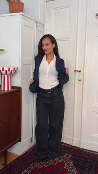 Γυναίκα με μακριά σκούρα μαλλιά που φοράει λευκό μπλουζάκι henley, σκούρο μπλε σακάκι και μαύρο τζιν με φαρδιά μπατζάκια, στέκεται μέσα σε κλειστό χώρο κοντά σε ντουλάπι και κλειστή πόρτα.