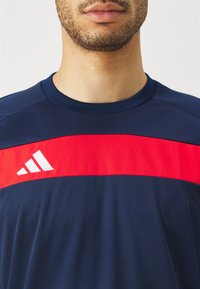 Navy athletic shirt featuring a horizontal red ribbed band across the chest with a white logo. Soft, smooth fabric; crew neck design.